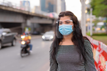 Fototapeta premium Young Indian woman with mask and face shield thinking in the city streets