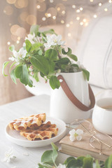 A bouquet of branches of a blossoming apple tree stands in a white vase on the table. Viennese waffles on a plate. Coffee in a white cup.