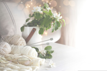 Knitting, knitting needles, yarn and glasses lie on a white wooden table. Surrounded by branches of a blossoming apple tree. A bouquet of branches is in a white vase.