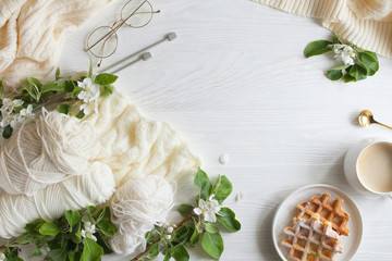 Knitting, knitting needles, yarn, coffee and waffles stand on a white wooden background. Surrounded by branches of a blossoming apple tree. Flatlay.
