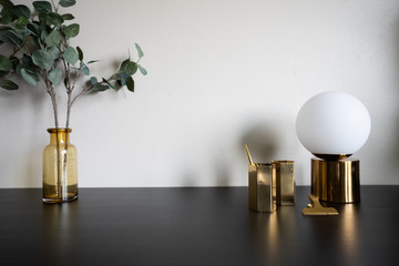 Bedroom working corner decorated with hexagon gold stainless vase ,gold circular lamp and artificial plant in glass vase on black working table with beige wall in the background /apartment interior