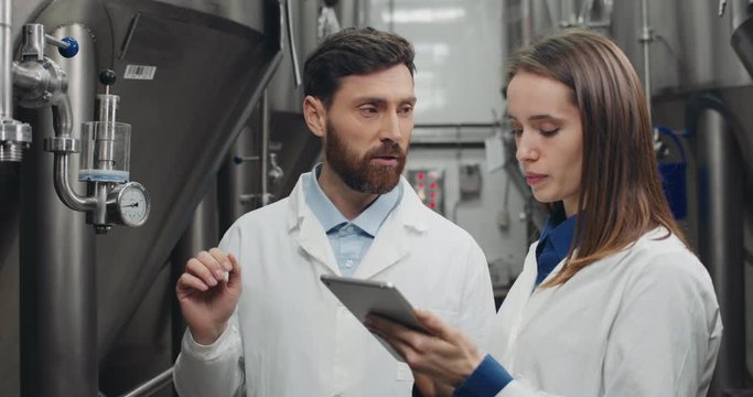 Male and female process engeneers looking at equipment gauge and talking. Man and woman in white lab coats entering data and using tablet while standing near automatic beverage machine