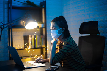 Asian woman wearing protective face mask working on a laptop overtime at home. WFH. Work from home for avoid the Coronavirus COVID 19 concept.