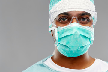 Fototapeta na wymiar medicine, surgery and people concept - indian male doctor or surgeon in mask, goggles and protective wear over grey background