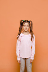 A girl with two ponytails stands on a pink background in daily clothes