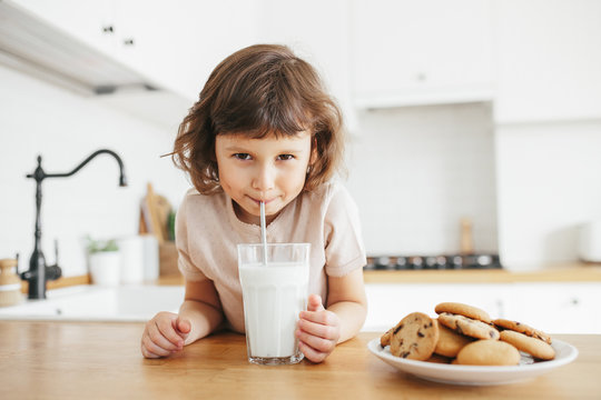 Cute toddler girl drinking milk with steel straw from glass and eating cookies