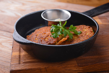 Delicious potato pancakes with sour cream served in a frying pan on wooden board over light rustic wooden table.
