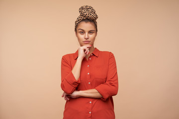 Pensive young pretty brunette lady squinting her eyes while looking thoughtfully at camera and holding chin with raised hand, isolated over beige background