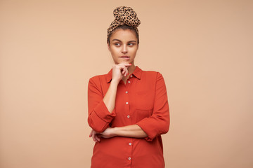 Studio shot of young thoughtful brown haired female with headband keeping raised hand on her chin and biting pensively underlip while posing over beige background