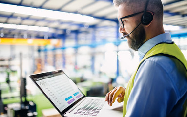 Side view of technician or engineer with headset and laptop standing in industrial factory.
