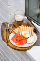 Toasted bread, salted salmon and poached egg with cream cheese and cup of coffee on white windowsill.
