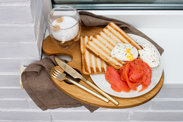 Toasted bread, salted salmon and poached egg with cream cheese and cup of coffee on white windowsill.