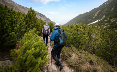 Two young men walk down a path with sticks and backpacks at the mountain. Hikers in Pirin mountain, Bulgaria. Beautiful spring green landscape