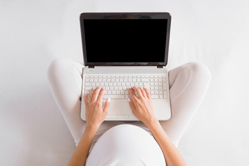 Young pregnant woman in white clothes sitting and using laptop in bed. Baby expectation. Homely atmosphere. Pregnancy information searching. Top view. Copy space. Empty place for text on black screen.