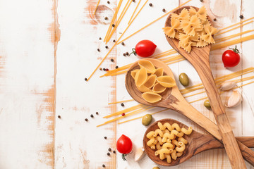 Different uncooked pasta with spices and vegetables on white wooden background