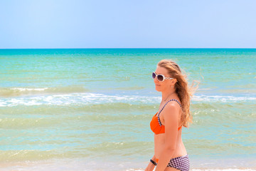 long-haired blonde in sunglasses in a swimsuit on the shore of the blue sea smiles, enjoying a summer sunny day, the beginning of the holiday season