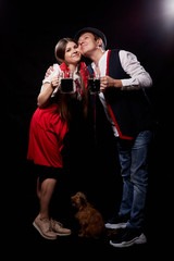 Funny Oktoberfest couple in national ethnic dress with mugs of beer, small shaggy dog kisssing on black background. Girl and boy with pet Brussels Griffon having fun with alcohol in Oktoberfest.