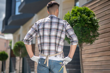 Dark-haired male in protective gloves and apron standing back to camera, pruning shears in his back pocket