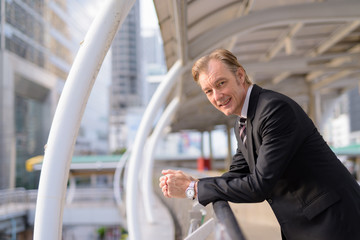 Happy mature handsome businessman in suit leaning at footbridge in the city