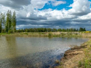 a fish pond, in the water of which there are colorful cumulus clouds and trees, there are recently dug gravel piles on the shore of the pond, the first spring greenery in nature