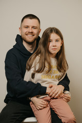 A young smiling father with a beard hugs strongly his cute daughter. A girl with long hair sitting on her father's lap. A happy family photo.