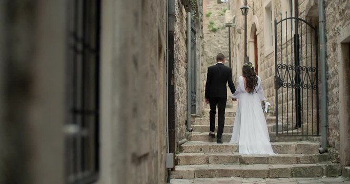 Back view of bride in beautiful white dress and groom walking in Europe old town, romantic wedding couple holding hands together in Montenegro.

