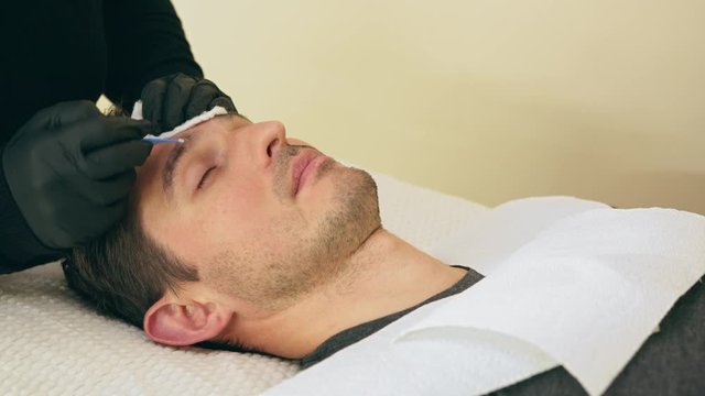 Close up of cosmetologist making permanent makeup. Microblading procedure. High quality close-up 4k slow motion video of a man laying on a couch during the esthetician brow microblading treatment.