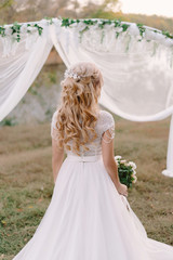 Beautiful tender happy bride with blonde curly hair and a head wreath standing in a sunset summer field, near wedding arch,enjoying herself, standing background to the camera