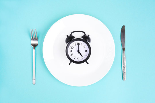Time to eat. Lunch time. View from above on white plate with alarm clock and knife and pork near on blue background. 5 p.m.