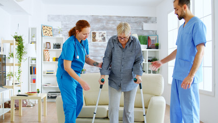 Obraz premium Senior woman with crutches getting help for female nurse and doctor to stand on her legs from the couch