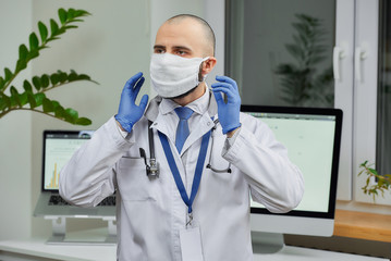 Fototapeta na wymiar A caucasian doctor correcting a protective face mask to avoid the spread coronavirus (COVID-19) in his office. A bald physician with a beard preparing to examine a patient.