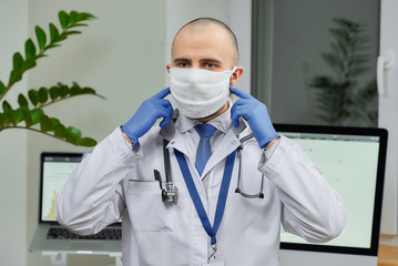 Fototapeta na wymiar A caucasian doctor correcting a protective face mask to avoid the spread coronavirus (COVID-19) in his office. A physician with a beard preparing to examine a patient.