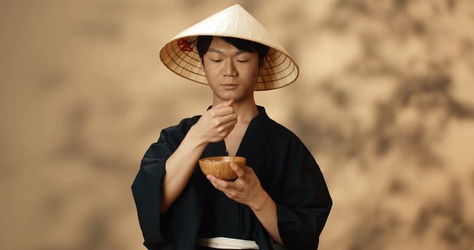 Portrait of handsome young Asian man in traditional clothes and conus hat holding plate with rice and pouring seeds from hand. Chinese guy with bowl of grain smiling to camera cheerfully.