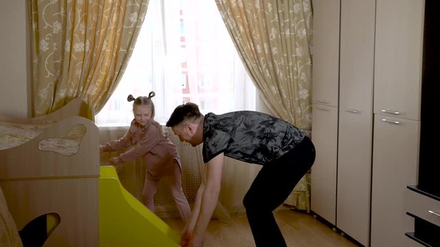 Enjoying old dad play by moving a ladder on wheels with a joyful little girl in a bright room in fashionable clothes. On against a bright window.