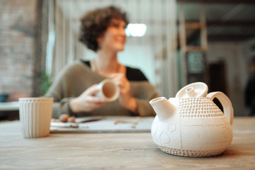 Clay teapot in the front, young woman on the background. Working in pottery.