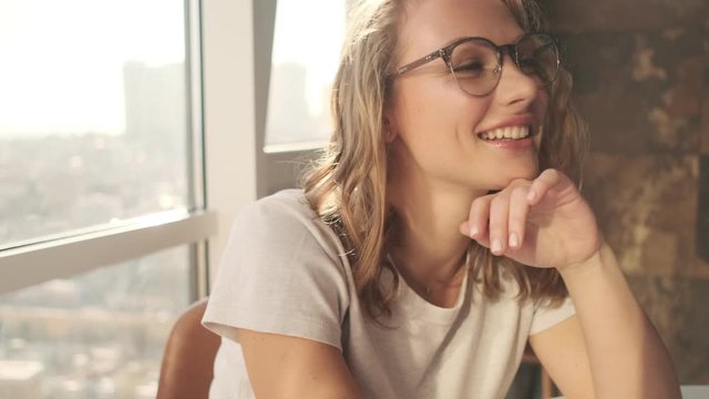 Young optimistic woman indoors at home wearing eyeglasses near window