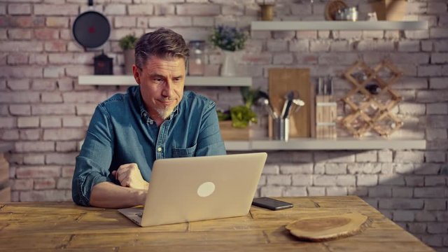 Man working from home on laptop computer, sitting at table in kitchen, looking busy.