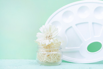 Working space. White flower in a glass jar with natural filling on a light green background. View from above. Copy space - concept art, entertainment, hobby