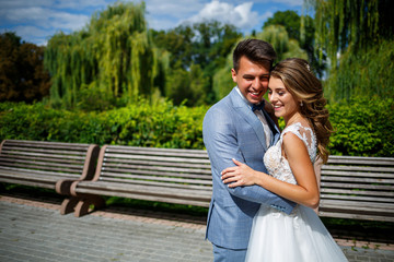 Naklejka premium Young stylish guy in a suit the groom and the bride beautiful girl in a white dress with a train walk in the park on their wedding day
