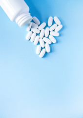 Pills of vitamin in the shape of heart with the opened white plastic container on soft blue background.
