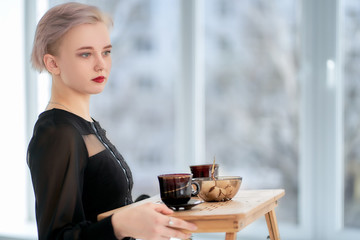 A cute woman in a black dress and a short haircut carries a tray with tea mugs.