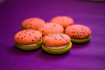 Multi-colored green and pink, bright cookies with two halves dressed with filling and sprinkled with sesame seeds on a bright purple color background.