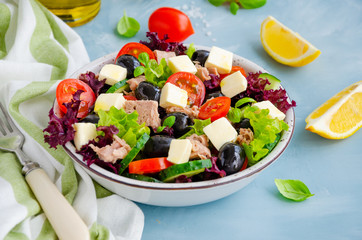 Salad with tuna, fresh vegetables, olives and feta cheese in a bowl on a light blue concrete background. Healthy food. Horizontal orientation.