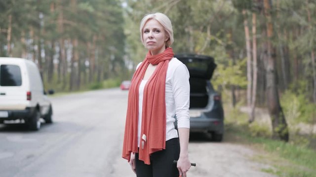 Side View Of Adult Blond Woman Standing Next To Broken Car With Wrench And Waiting For Tow Truck. Portrait Of Sad Caucasian Female Driver Having Traffic Accident On Countryside Road.