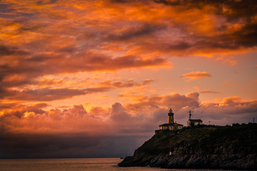 Hermoso atardecer sobre la ría con la silueta del faro sobre la montaña con colores cálidos en las nubes 