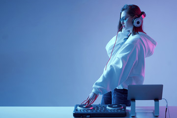 Cool young girl DJ mixes music on a mixing console and laptop, in stylish clothes, glasses on a...