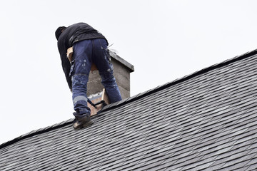 Roofer construction worker repairing chimney on grey slate shingles roof of domestic house, sky background with copy space.
