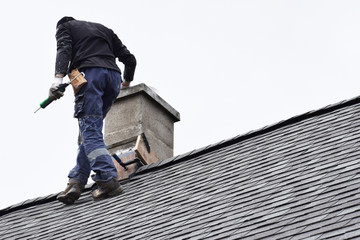 Roofer construction worker repairing chimney on grey slate shingles roof of domestic house, sky...