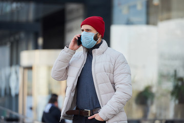 A caucasian man wearing a medical face mask to avoid the spread coronavirus (COVID-19) in the center of the city. A guy with a surgical mask on the face calling on the phone.