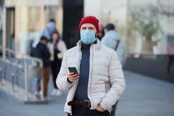 A caucasian man wearing a medical face mask to avoid the spread coronavirus (COVID-19). A guy with a surgical mask on the face holding a smartphone in the center of the city.